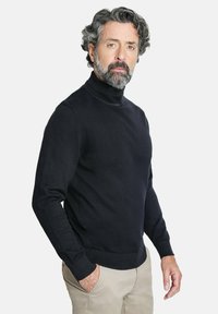 Suéter negro de cuello alto con un diseño ajustado, fabricado en una suave tela de punto, con puños y dobladillo acanalados. Combinado con pantalones beiges.