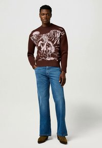 Rjavi pleteni pulover s rožnato planinsko obliko, kombiniran z svetlo modrimi razširjenimi kavbojkami in rjavimi antilop čevlji.