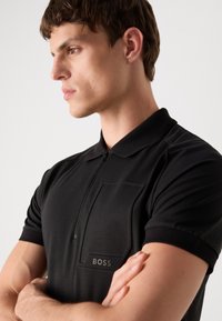 Μαύρο πόλο μπλουζάκι με κολάρο που κλείνει με φερμουάρ, κοντά μανίκια και τσέπη στο στήθος. Διαθέτει διακριτικό ανάγλυφο λογότυπο 'BOSS' στην τσέπη.