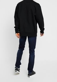 Sudadera negra con cuello redondo, combinada con jeans de corte ajustado en azul oscuro. Zapatillas negras con suelas y detalles blancos completan el atuendo.