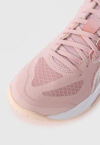 Κοντινή λήψη αθλητικού παπουτσιού σε ανοιχτό ροζ χρώμα που δείχνει υφασμάτινες επιφάνειες με υφή, κορδόνια και μπεζ σόλα σε λευκό φόντο.