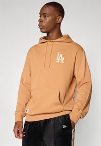 Hoodie beige avec une poche avant et un logo "LA" en blanc. Tissu doux, capuche avec cordon de serrage et poignets côtelés. Porté avec un pantalon noir arborant des logos.