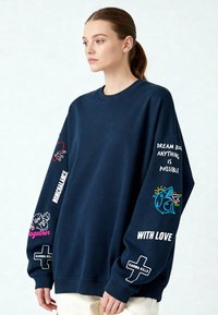 Jūras zila lielizmēra krekls ar garām piedurknēm. Tam ir krāsaini izšuvumi: frāzes un dizaini uz piedurknēm, ieskaitot sirdis un haizivi.