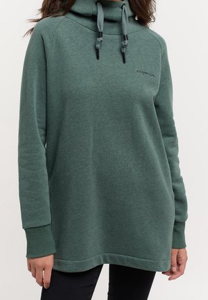 Groene pullover met lange mouwen en capuchon met dikke trekkoorden, gedragen door een persoon met een donkere broek, afgebeeld van schouders tot halverwege de dijen.
