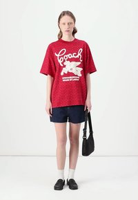 T-shirt curto vermelho de manga curta com texto gráfico branco e um logótipo, combinado com calções de ganga escuros. O modelo segura uma pequena mala preta de ombro.