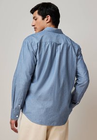 Uomo che indossa una camicia a maniche lunghe blu con strisce verticali bianche, di spalle, abbinata a pantaloni beige chiaro su uno sfondo neutro.