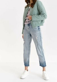 Frau in einer hellgrünen gesteppte Jacke, pinkfarbenem grafischen T-Shirt, ausgefransten blauen Jeans und weißen Turnschuhen, die vor einem weißen Hintergrund steht.