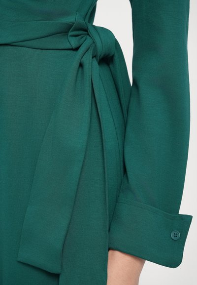 Zöld ruha sima anyaggal, oldalt megkötővel és gombos mandzsettával az ujjon. Egyszerű kialakítás és elegáns sziluett.