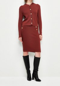 Rdzawy prążkowany sweter zapinany na guziki z złotymi guzikami, zestawiony z pasującą prążkowaną spódnicą z detalami bocznych guzików oraz czarnymi skórzanymi kozakami za kolano.