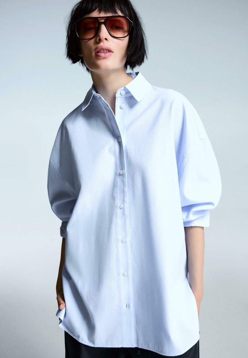Jasnoniebieska, oversize koszula z guzikami, z wyraźnym kołnierzykiem, obszerne rękawy i gładka tekstura, w połączeniu z dużymi okrągłymi okularami przeciwsłonecznymi.