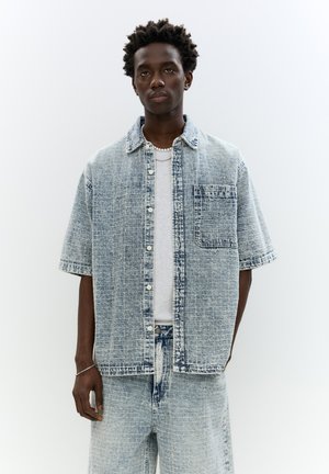 Short-sleeve denim shirt in light blue with a textured pattern, buttons down the front, and a chest pocket. Worn over a white T-shirt.