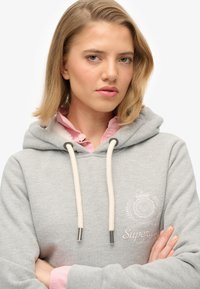 Sudadera gris con bolsillo frontal y cordones; presenta detalles de logotipo en blanco y acentos en rosa claro en el cuello. Textura de tela suave.