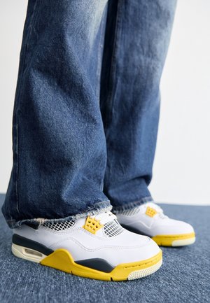Personne portant un jean en denim bleu et des baskets blanches avec des détails jaunes et noirs, debout sur un tapis bleu.