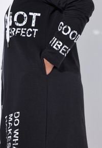 Zwarte hoodie jurk van zacht materiaal, met witte tekstontwerpen, een voorzak en een ritsluiting met een trekkoord capuchon.