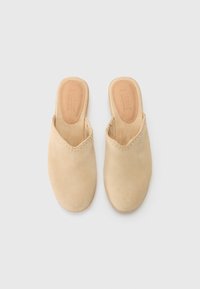 Beige mocka slip-on mules med en mjuk textur och fransade kanter. Läderinnersulan har en präglad logotyp. Ingen häl. Minimalistisk design.