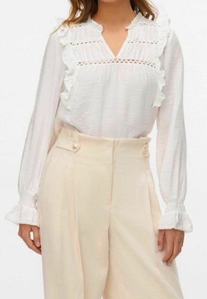 Femme portant un chemisier blanc à manches longues avec volants, rentré dans un pantalon beige taille haute à jambes larges avec des détails à boutons.