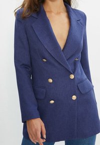 Blazer croisé bleu marine en tissu texturé, doté de boutons dorés, de revers à encoche et de deux poches avant.