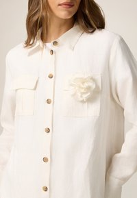 Camicia bianca di lino con colletto, due tasche sul petto e un fiore decorativo in tessuto sulla tasca sinistra. Presenta bottoni in tono dorato.