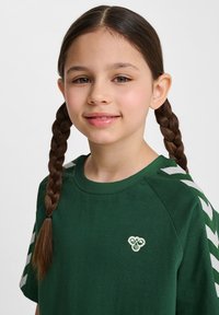 Camiseta de algodón verde con cuello redondo, que presenta un pequeño logo y rayas en forma de cheurón blancas en las mangas. Textura suave y corte relajado.