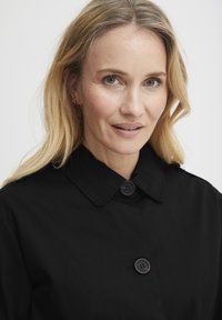 Melnas pogas mētelis ar apkakli, divām pogām un gludu tekstūru. Modelis ar viļņotiem blondiem matiem, demonstrējot dabiskas sejas iezīmes.