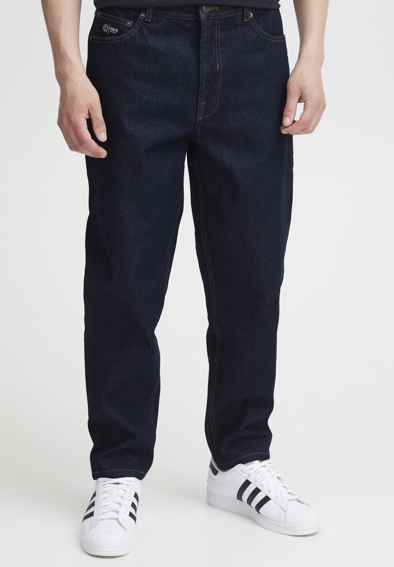 Temno modre denim hlače z ohlapno prileganjem, z oranžnim šivom in žepi. Združene z belimi supergami s črnimi črtami.