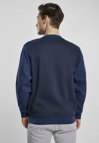 Mørkeblå hettegenser med rund hals, ribbestrikkede ermer og kant. Har en glatt tekstur og tettsittende design, vist fra baksiden.