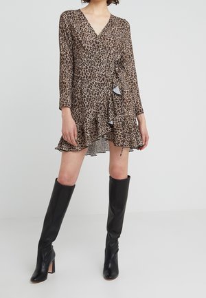 Femme portant une robe portefeuille marron à imprimé léopard avec des détails à volants et des bottes noires montantes à talons, debout sur un sol blanc uni.