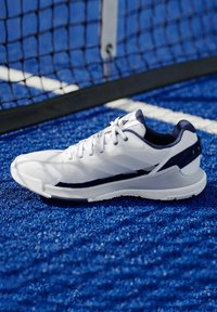 Bílé sportovní boty s texturovaným svrškem, námořnickými akcenty a měkkou podrážkou, umístěné na modrém syntetickém trávníku.