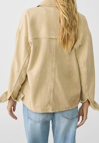 Veste en denim beige au fit oversize, avec des coutures visibles et des fentes latérales. Elle présente un dos avec un empiècement et une petite étiquette de marque sur l'ourlet inférieur.