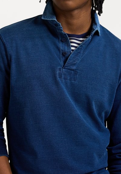Blå tröja med en krage i denimstil, tre knappar fram och en framficka. Mjuk, strukturerad tyg med en avslappnad passform. Randig skjorta under.