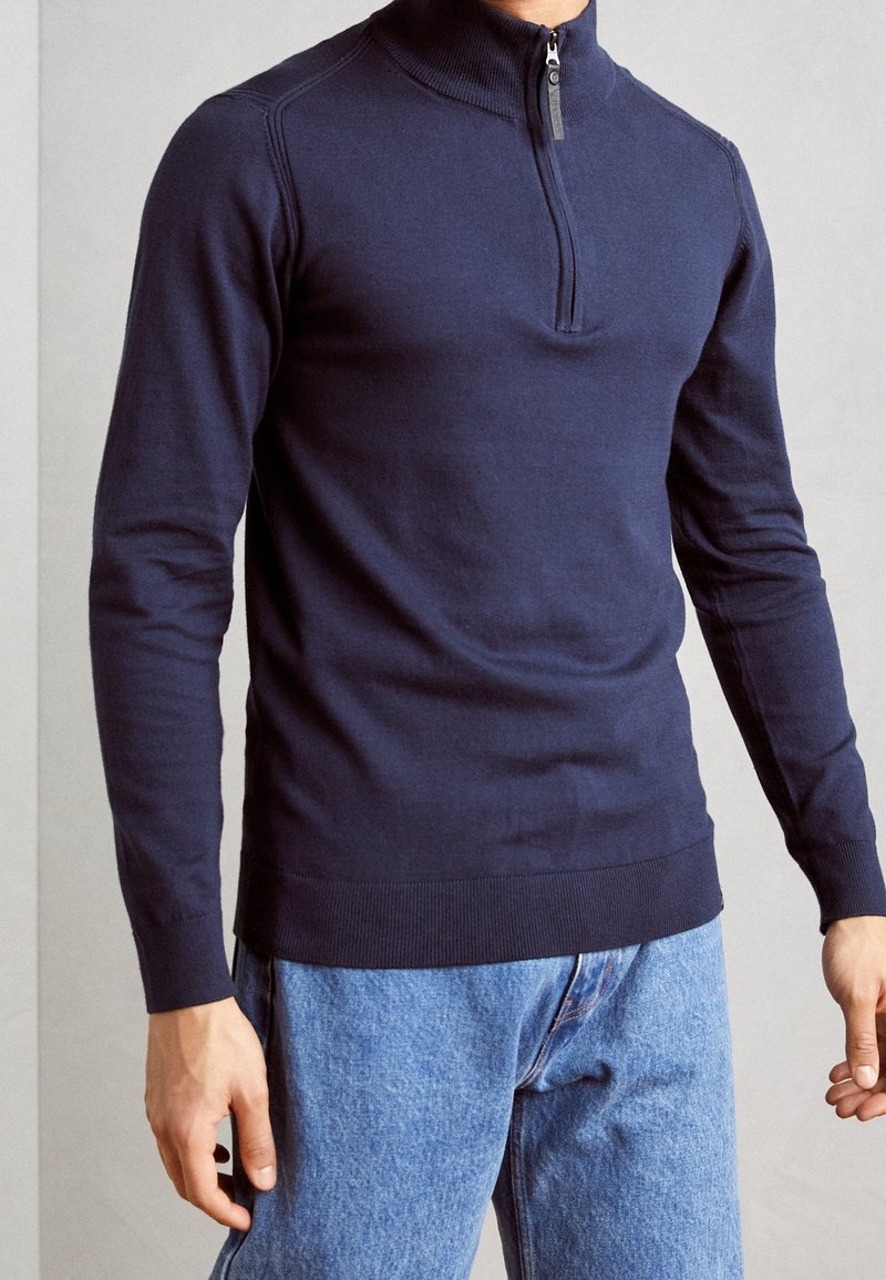 Uomo che indossa un maglione blu navy con zip al collo e jeans azzurri, in piedi con le braccia rilassate contro uno sfondo semplice.