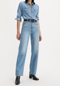 Camisa de denim azul claro con estilo abotonado, dos bolsillos en el pecho. Vaqueros de tiro alto azul claro, corte de pierna ancha, combinados con botines negros de tacón.