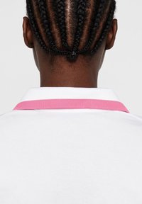 Bakifrån av en person med tätt flätat hår i cornrows, iklädd en vit skjorta med krage och en rosa rand på kragen.