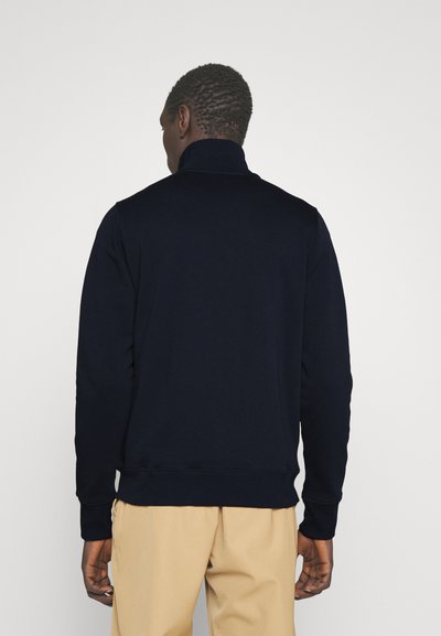 Sweatshirt bleu marine avec un col montant, des poignets et un ourlet côtelés ; fabriqué en tissu doux. Porté avec un pantalon beige. Vue de dos.