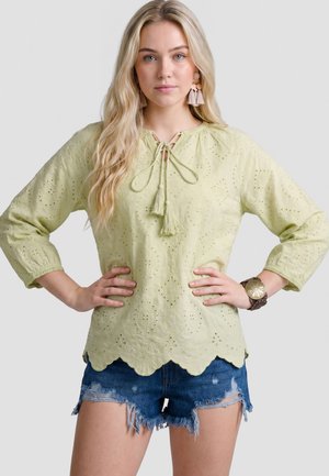 Jonge vrouw met lang blond haar draagt een lichtgroene blouse met oogjes en kwastjes en blauwe versleten spijkershorts, handen in de heupen.