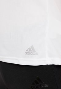 Camiseta deportiva blanca con una tela texturizada, que presenta un sutil logo gris de Adidas cerca del dobladillo, combinada con pantalones deportivos negros que exhiben el mismo logo.