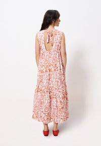 Blumiges Midikleid in Rosa- und Orangetönen, mit einem Rückenausschnitt und einer Schleifendetail und einem gestuften Rockdesign. Rote Schuhe sichtbar.