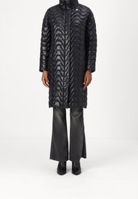 Abrigo inflado negro con un patrón de ondas, cuello alto y cremallera frontal. Lleva puesto sobre unos vaqueros grises de pierna ancha y botines negros.