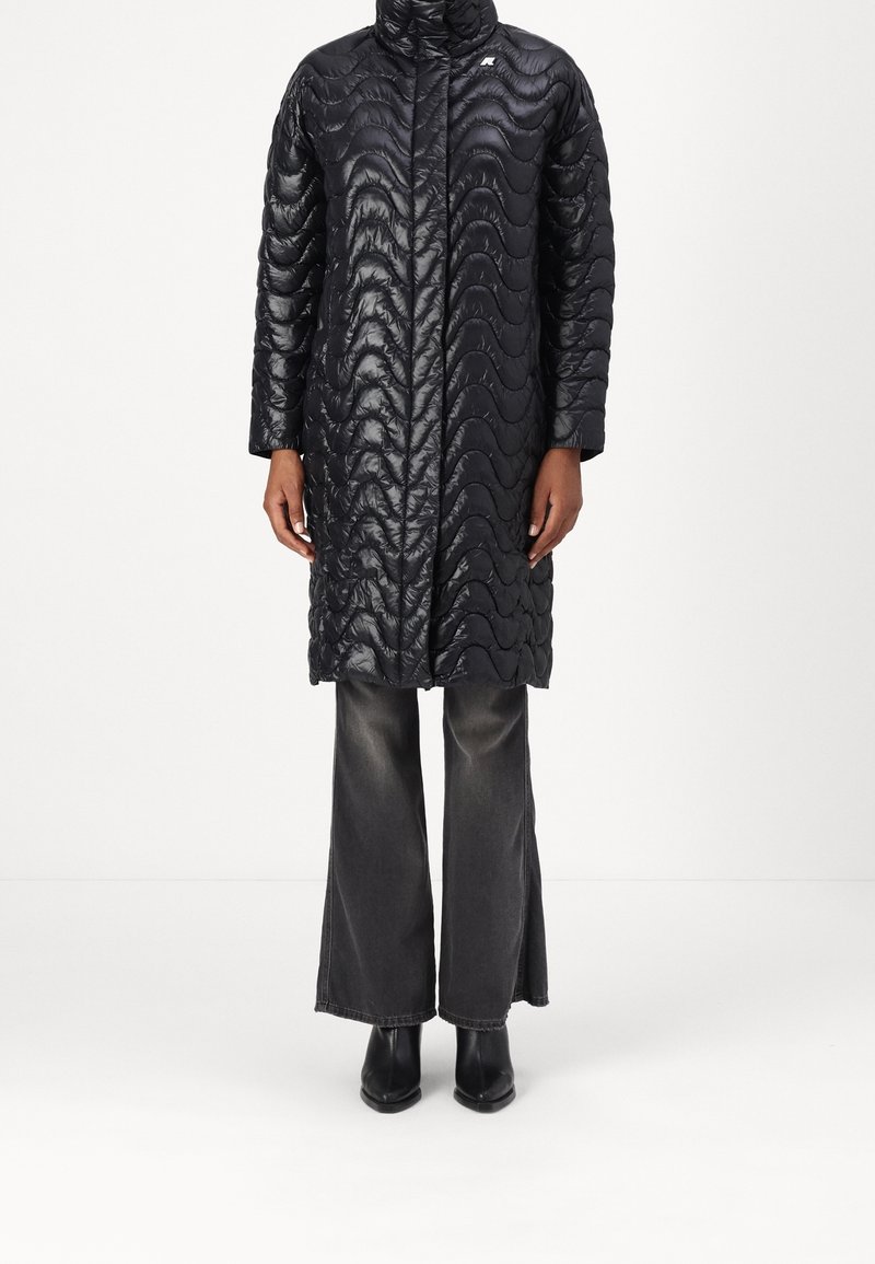 Abrigo inflado negro con un patrón de ondas, cuello alto y cremallera frontal. Lleva puesto sobre unos vaqueros grises de pierna ancha y botines negros.