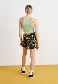 Grün gestreiftes Tanktop, kombiniert mit schwarzen Blumen-Shorts mit lebhaften Pflanzenmustern, getragen mit weißen Socken und schwarzen Schuhen.