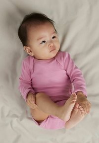 Langærmet lyserød heldragt på en spædbarn, der sidder med benene krøllet, mod en lys beige baggrund, der viser blødt stof og tryklås.