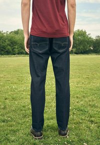 Persoon staat op een grasveld, draagt donkerblauwe jeans en een rood T-shirt, met de rug naar de camera en handen ontspannen langs het lichaam.