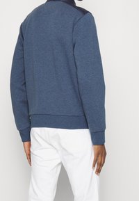 Blauwe trui met een zachte textuur, voorzien van een donkere kraag en accenten op de schouders, geribbelde manchetten en een ontspannen pasvorm, gedragen met witte broek.