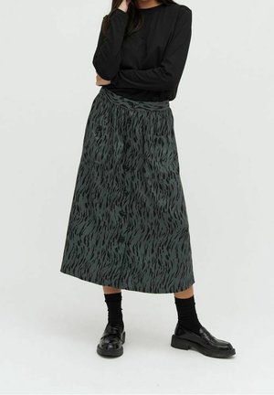 Frau trägt einen langen schwarzen gemusterten Rock mit vorderen Knöpfen, ein schwarzes langärmliges Oberteil, schwarze Socken und schwarze Loafers und steht mit verschränkten Armen.