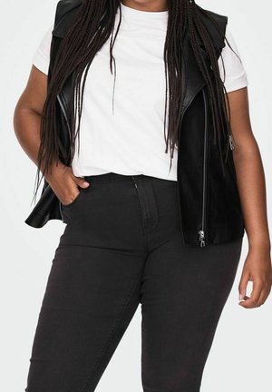 Kobieta z długimi warkoczami, ubrana w białą koszulkę, czarną skórzaną kamizelkę i czarne spodnie z wysokim stanem, ze zgiętą ręką w kieszeni, stojąca na białym tle.