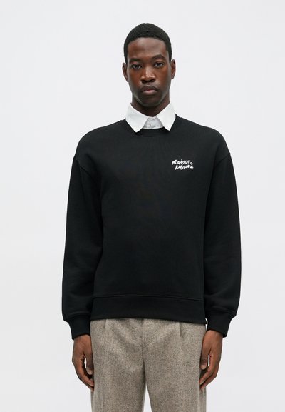 Čierna mikina s okrúhlym výstrihom a voľným strihom. Má biele logo na hrudi. Nosená cez bielu košieľu s golierom.