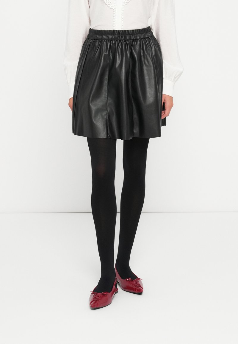 Jupe plissée en faux cuir noir avec taille élastique, portée sur des collants opaques noirs, associée à des chaussures rouges brillantes à bout pointu.