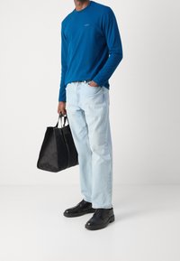 Blå långärmad skjorta, ljusblå jeans, svarta läderskor och en svart texturerad tygväska, hålls i vänster hand.