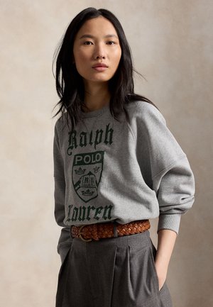 Ung kvinde med langt sort hår iført en grå Ralph Lauren sweatshirt, brun flettet bælte og grå plisserede bukser med hænderne i lommerne.