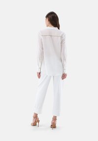 White long-sleeve shirt made of sheer fabric, featuring a buttoned back yoke, with tailored white trousers and beige high-heeled sandals.